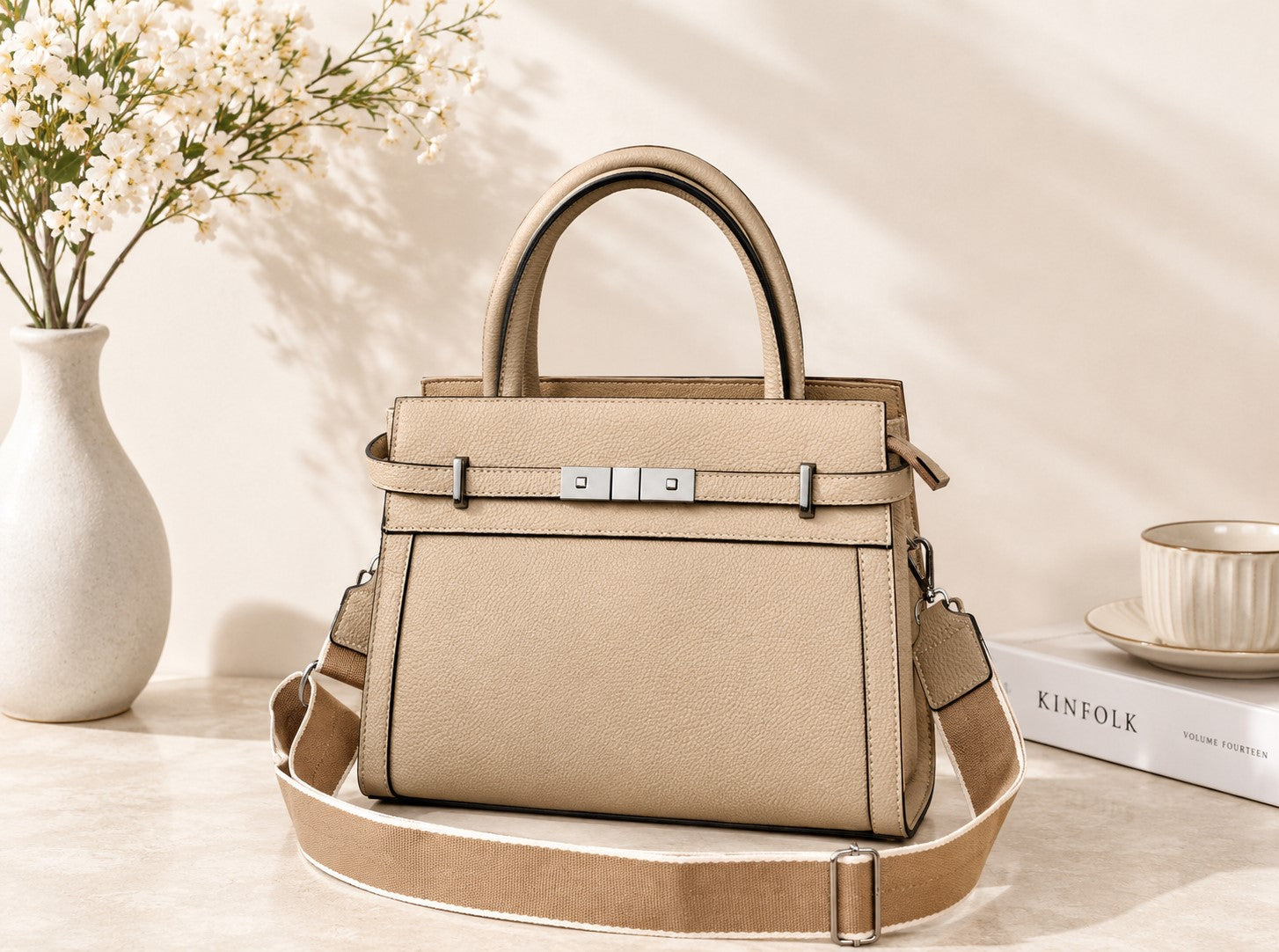 Beige handbag on a light surface with a vase of flowers and books in the background